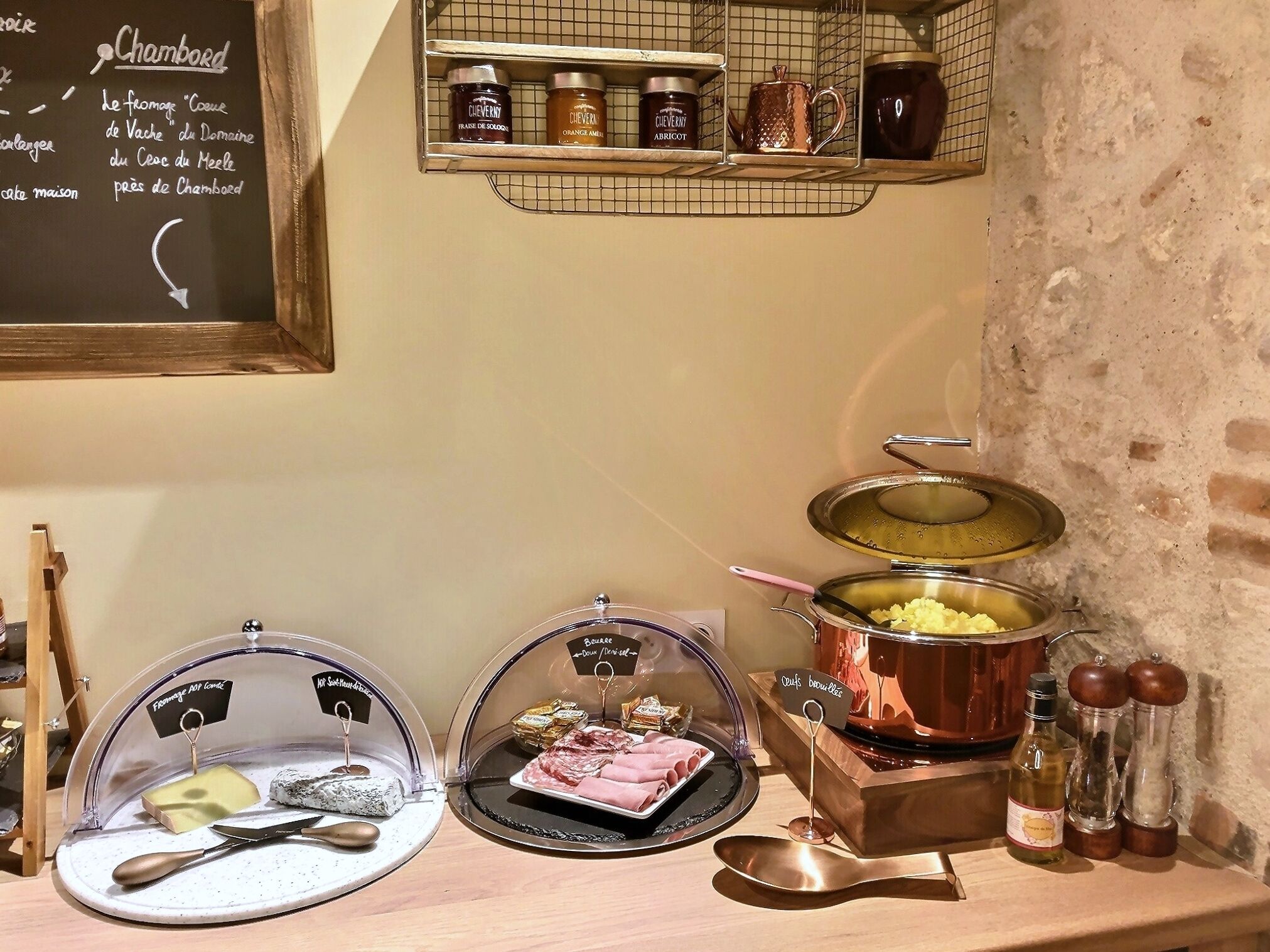 Una preparación para el desayuno con un menú en pizarra, frascos de mermelada, una olla de cobre y una presentación de quesos.