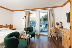 Relax in a spacious room with a large bed, armchair, and desk, offering a view of the surrounding buildings and trees.
