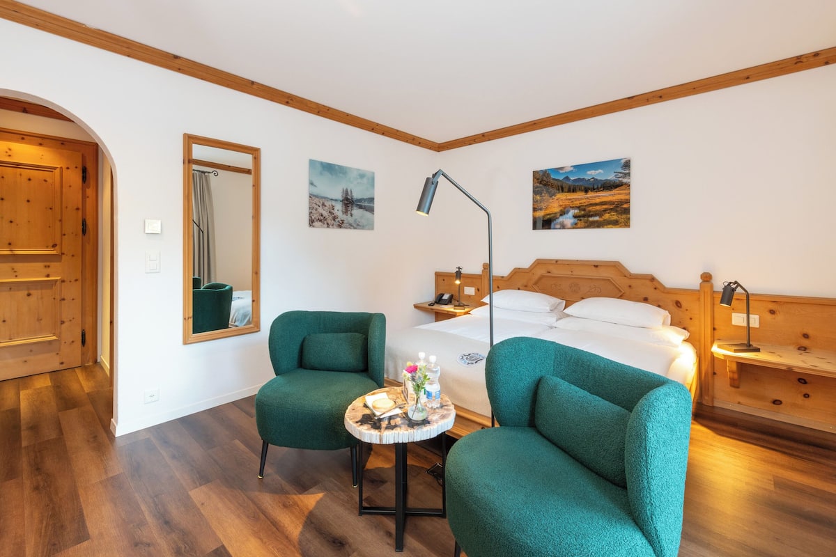 A room with a wooden bed, two green armchairs, a small round table with a book and flowers, and two wall-mounted lamps.