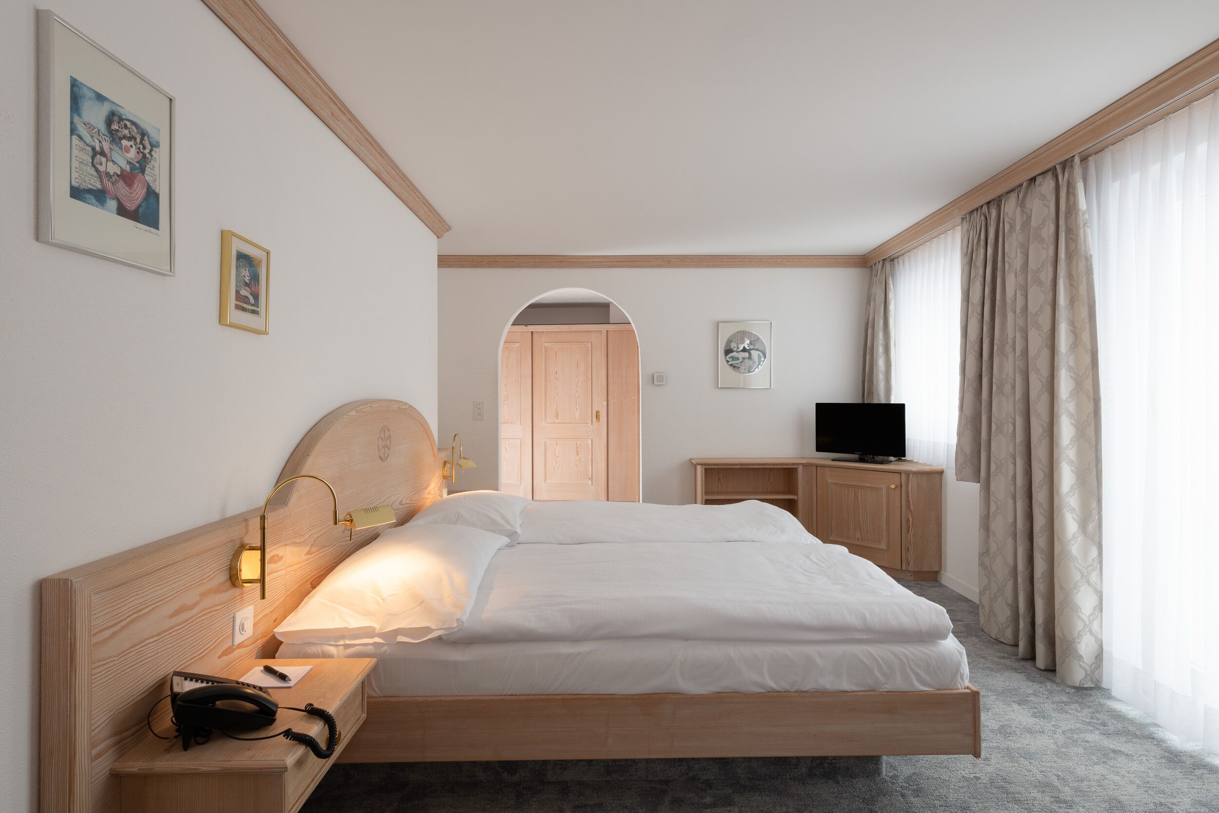 A hotel room with a wooden bed, a bedside table with a lamp, a telephone, and a television on a wooden cabinet.