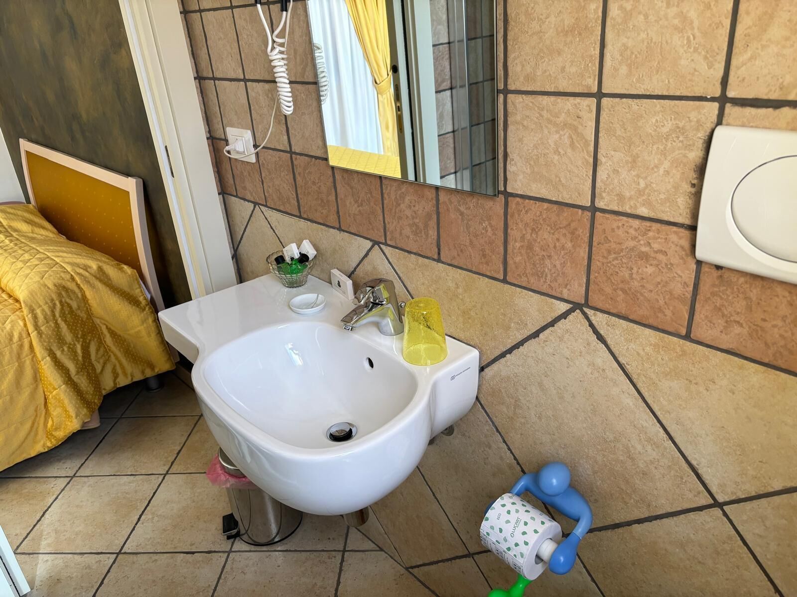 Une salle de bain avec un lavabo blanc, un miroir, des toilettes et un lit recouvert d’une couverture jaune.