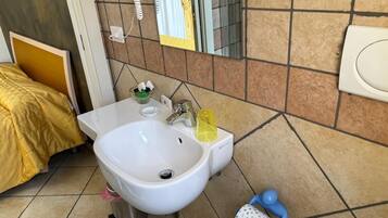 Une salle de bain avec un lavabo blanc, un miroir, des toilettes et un lit recouvert d’une couverture jaune.