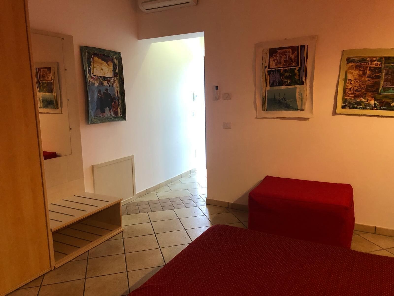 Une pièce avec un lit, un banc rouge, une étagère en bois et un couloir menant à une autre pièce.