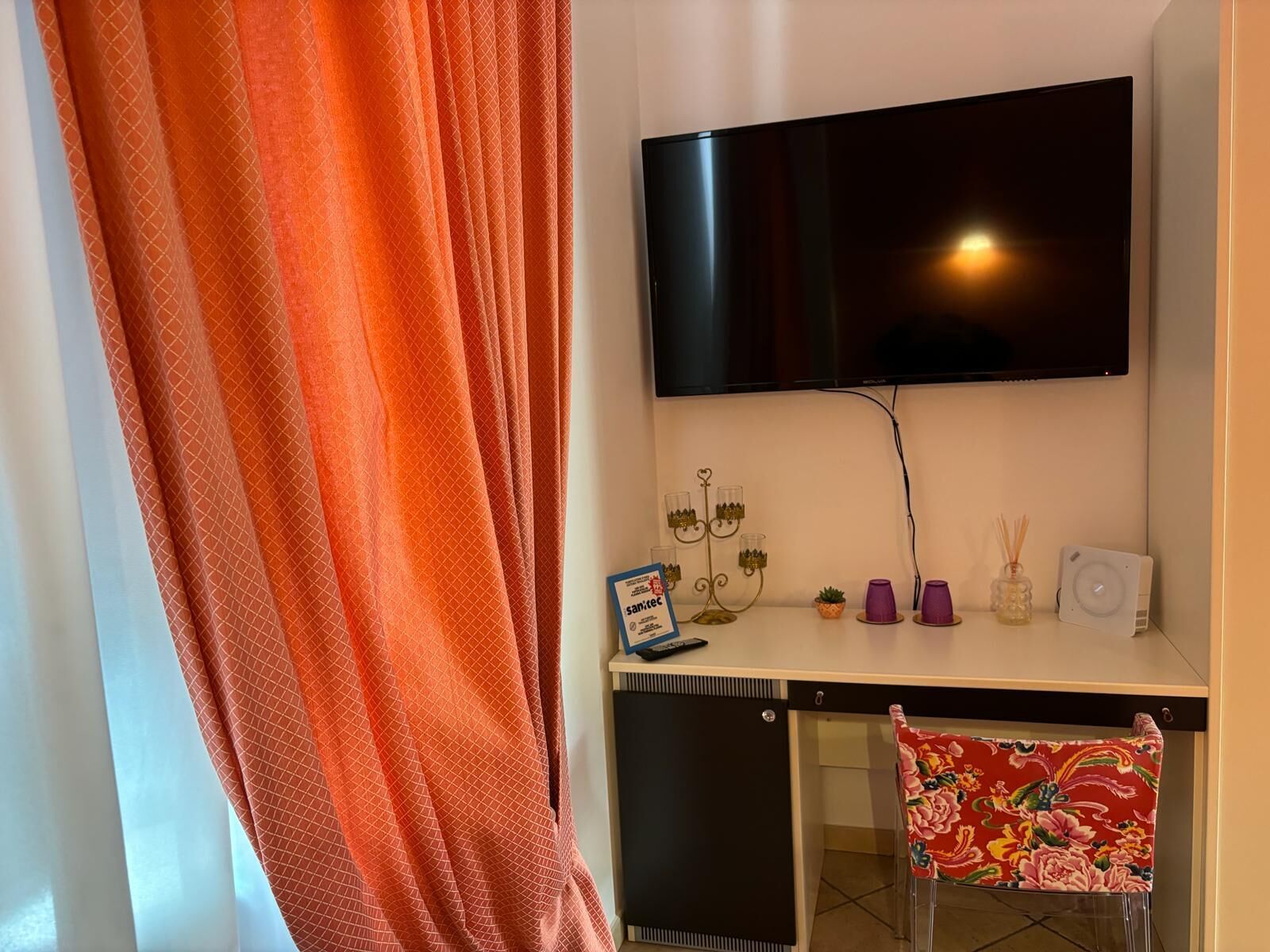 Una stanza con una TV a muro, una scrivania con un computer, una sedia e un tavolino con soprammobili.