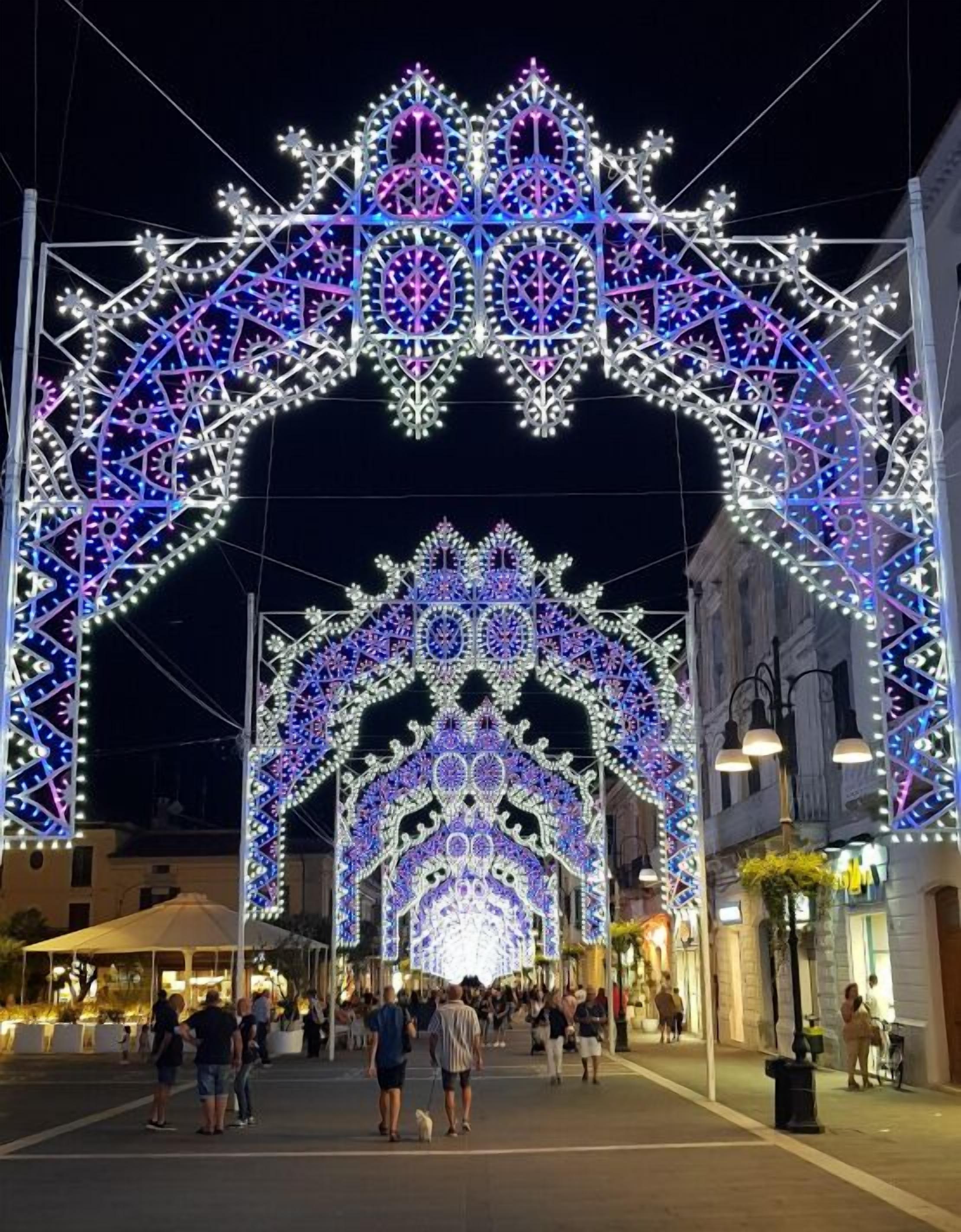 Un arco iluminado con diseños intrincados y luces, que conduce a una plaza pública con gente caminando y un edificio a la derecha.