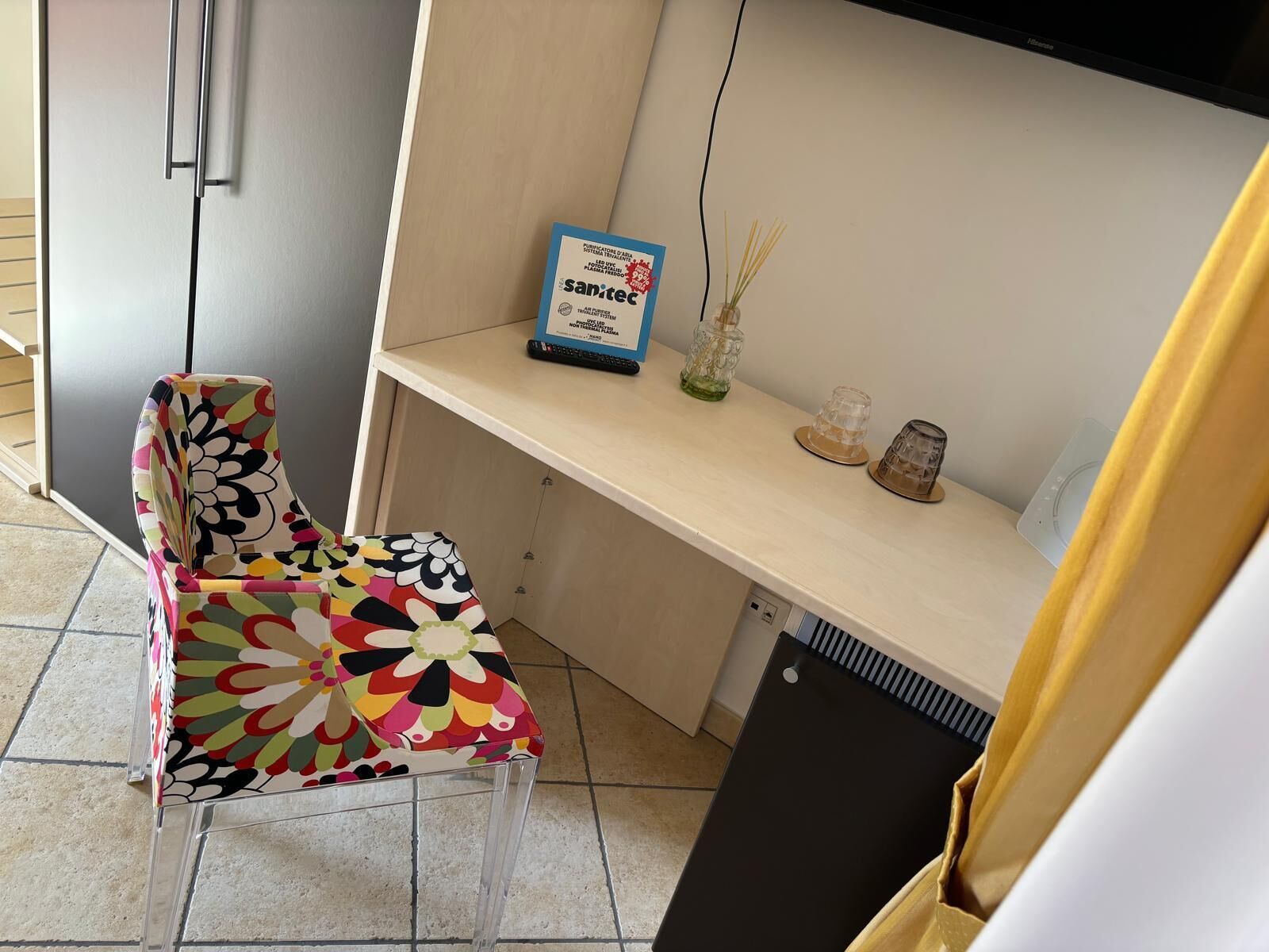 Una habitación pequeña y moderna con una silla colorida, un escritorio con un cartel azul y un pequeño jarrón con cañas.