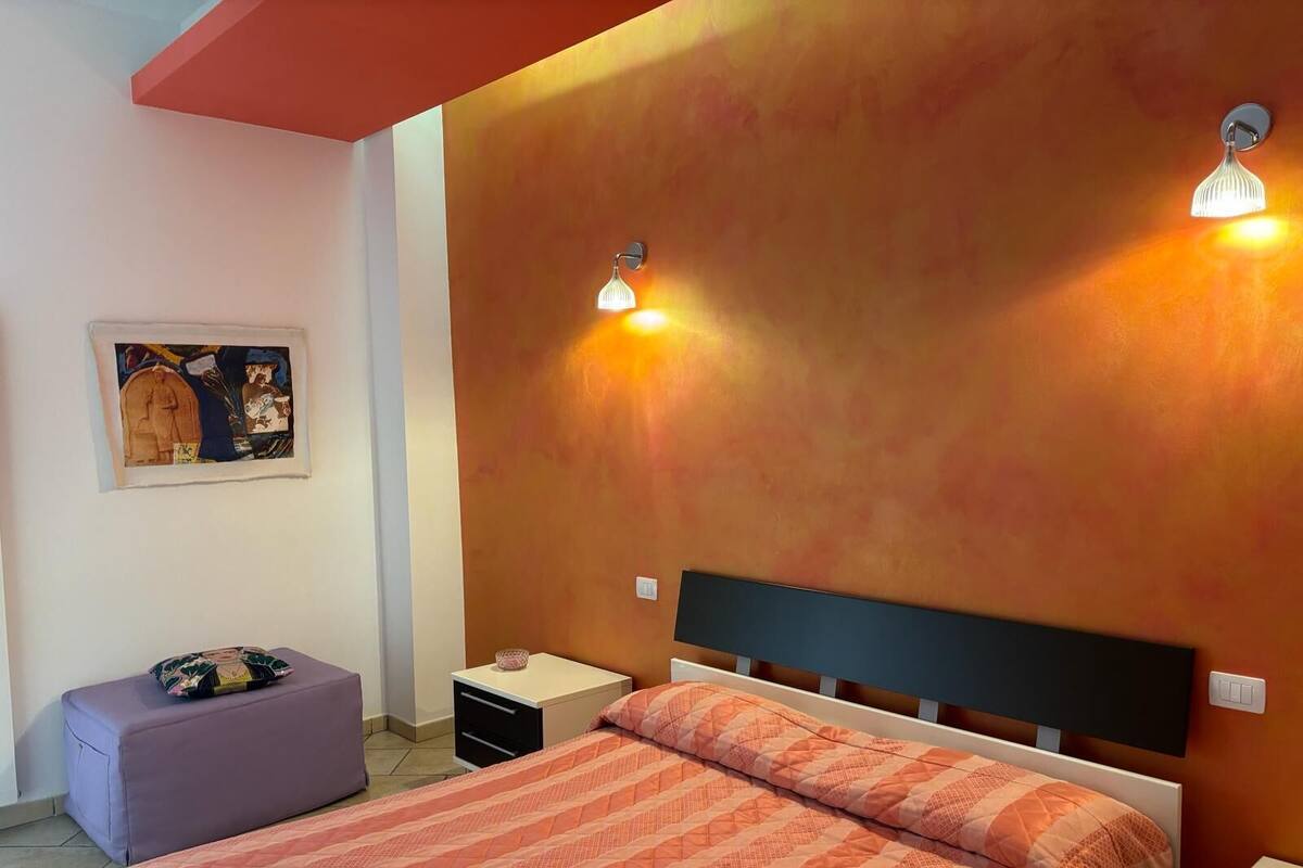 Ein Schlafzimmer mit einem Bett, einem Nachttisch, einem kleinen Tisch, einem violetten Hocker und einer Wandleuchte.