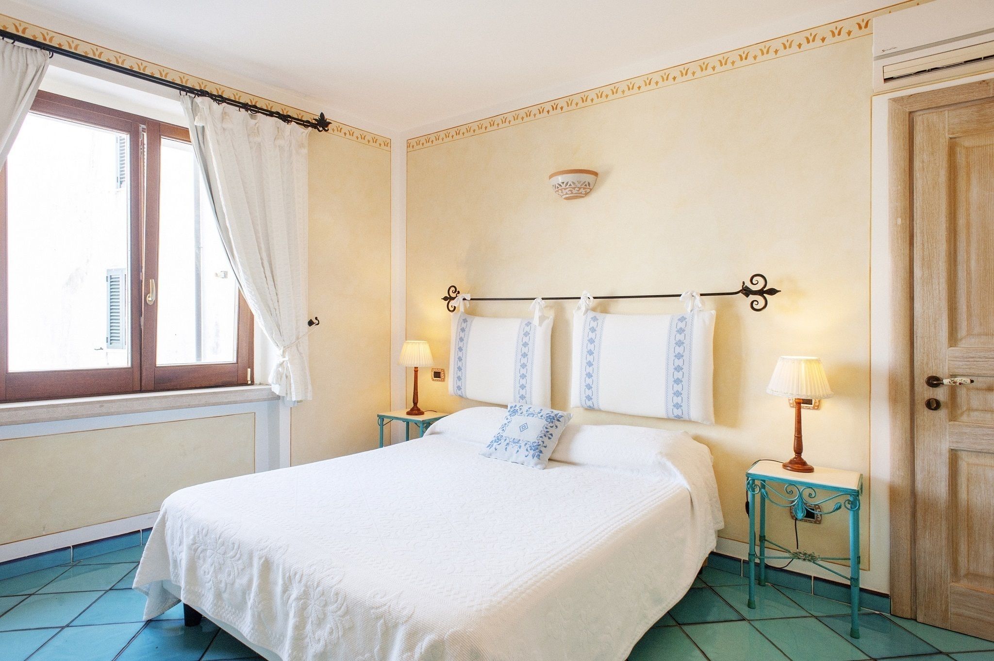 A bedroom with a bed, two bedside tables with lamps, a window with curtains, and a door.