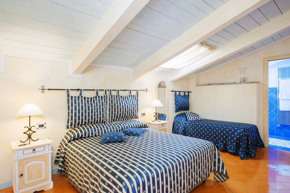 A bedroom with two beds, each with a blue and white striped bedspread, a bedside table with a lamp, and a small bedside table with a clock.