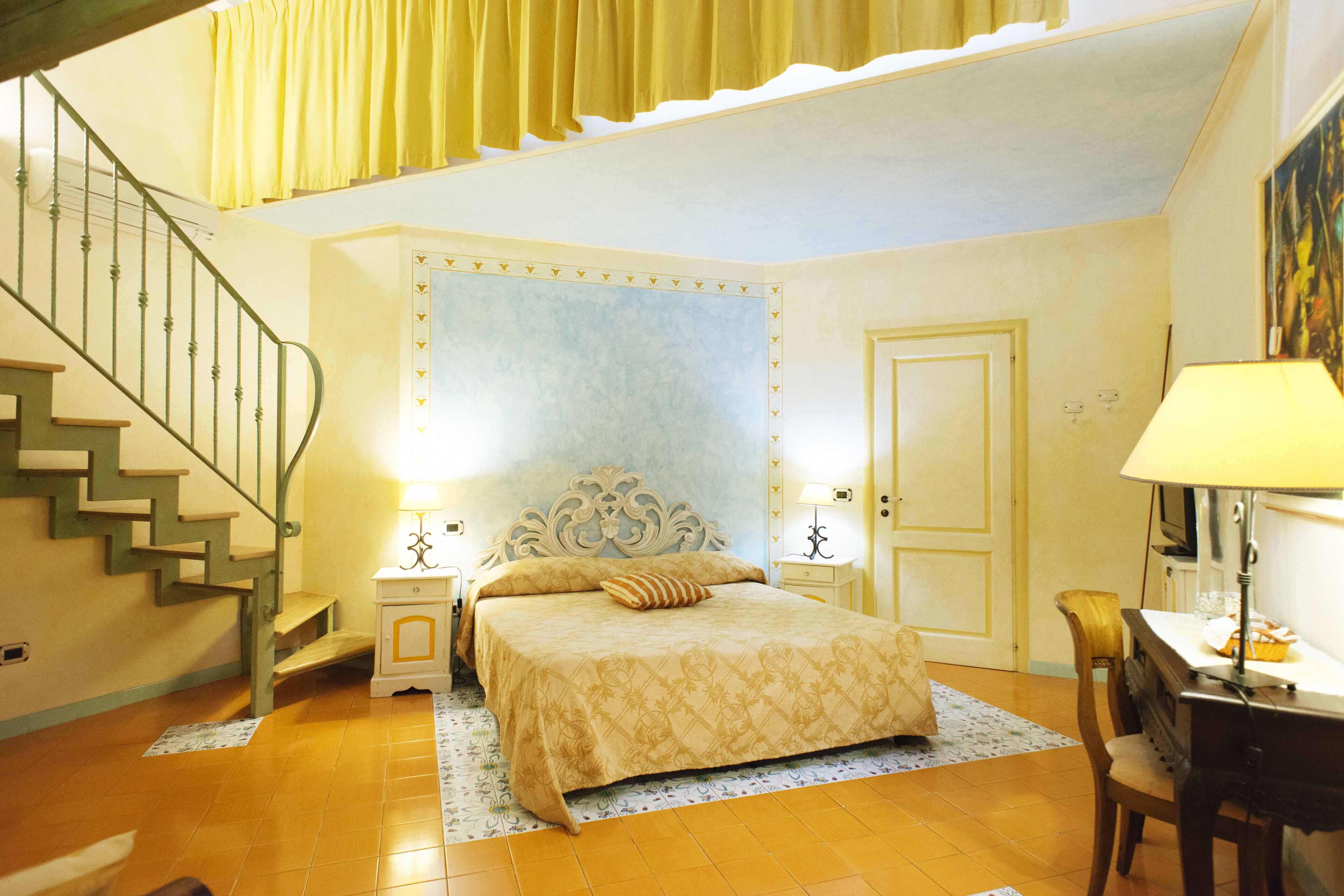 Ein Schlafzimmer mit einem Bett, einem Nachttisch, einer Lampe, einem Sessel, einem Schreibtisch, einem Gemälde, einer Treppe und einer Tür.