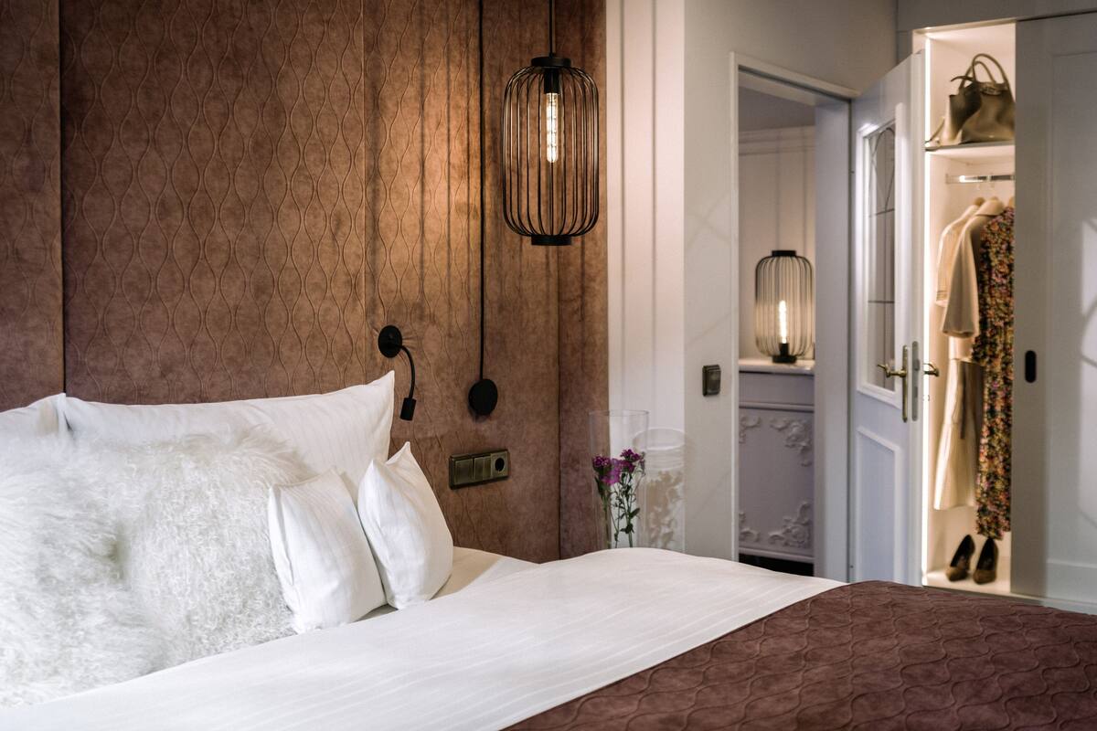 Un lit bien fait, avec du linge de lit blanc et une couvre-lit marron à motifs, un luminaire fixé au mur et une porte donnant sur un placard contenant un mannequin et des chaussures.