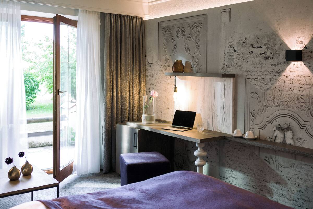Une chambre avec un lit, un bureau avec un ordinateur portable, un vase avec une fleur, et une vue sur la verdure à l’extérieur.
