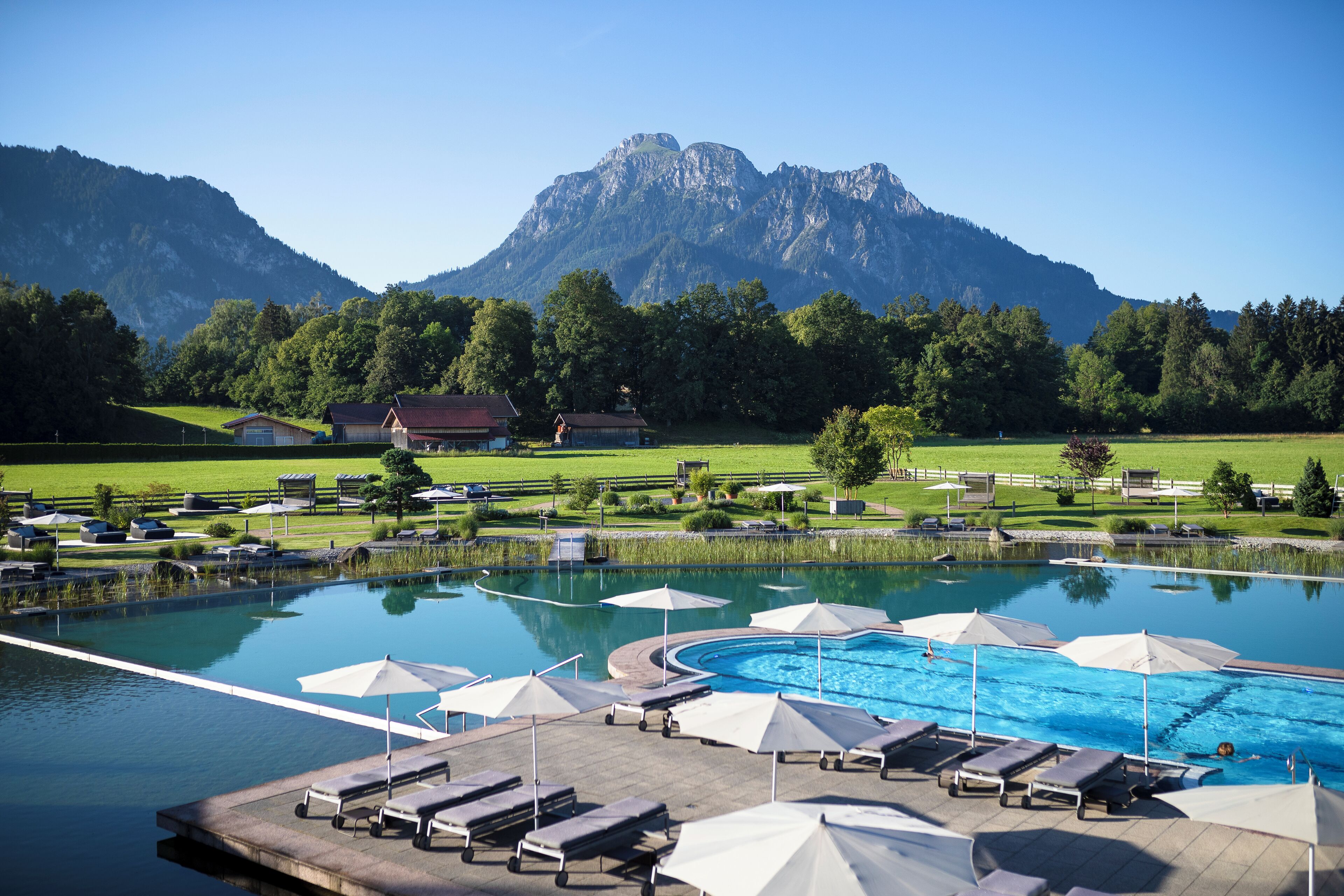 A swimming pool with lounge chairs and umbrellas, surrounded by a landscaped area with a mountain backdrop.