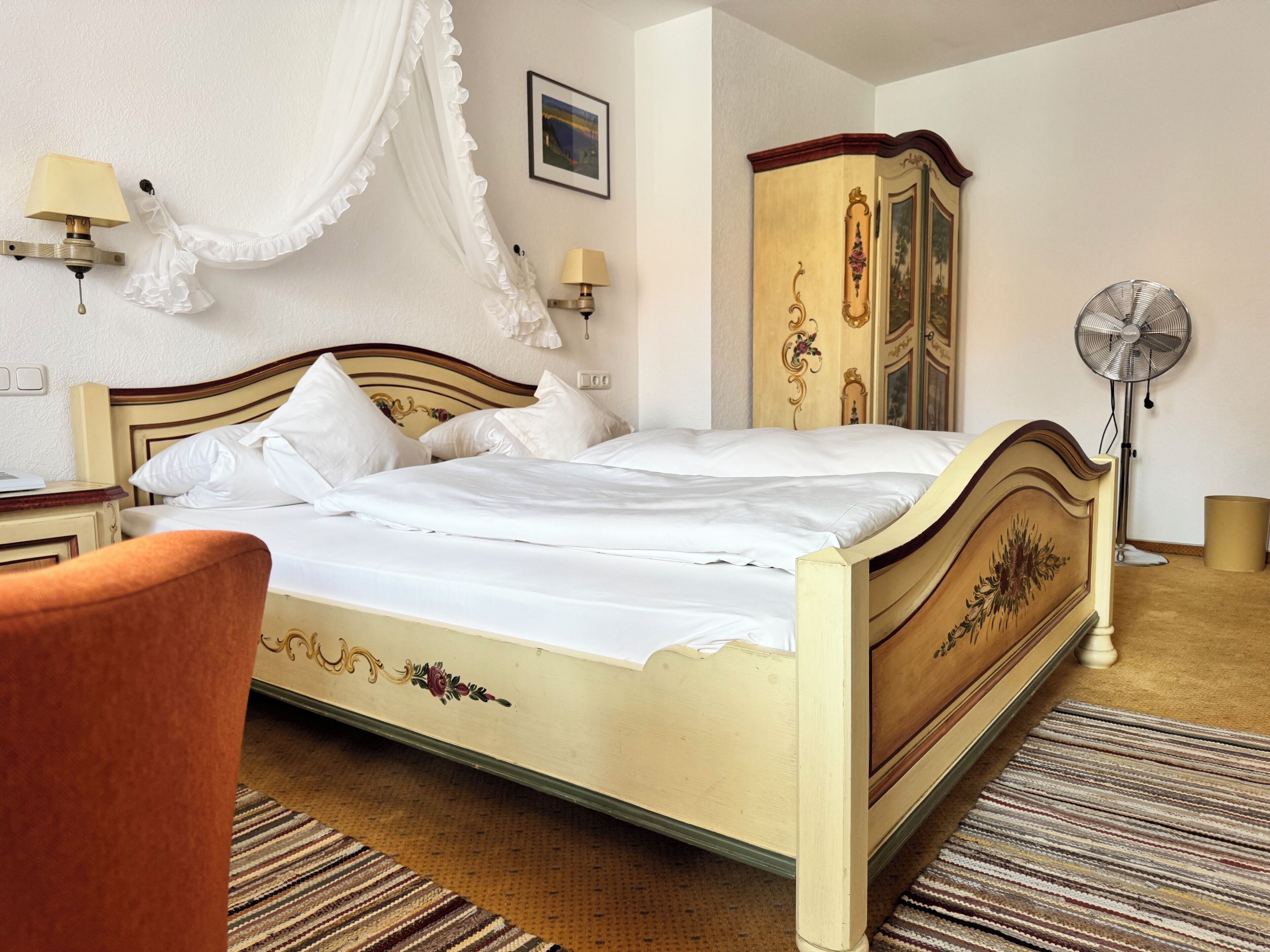 A hotel room with a bed, a chair, a wardrobe, a bedside table, a lamp, a picture on the wall, and a floor lamp.