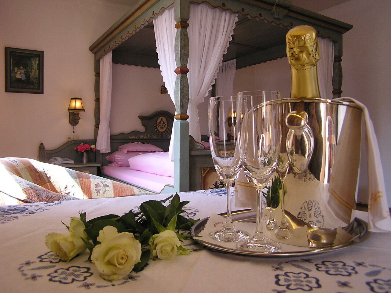 Un dormitorio con cama con dosel, una mesita de noche con lámpara y una bandeja con copas de champán y rosas.