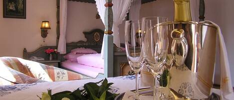Un dormitorio con cama con dosel, una mesita de noche con lámpara y una bandeja con copas de champán y rosas.