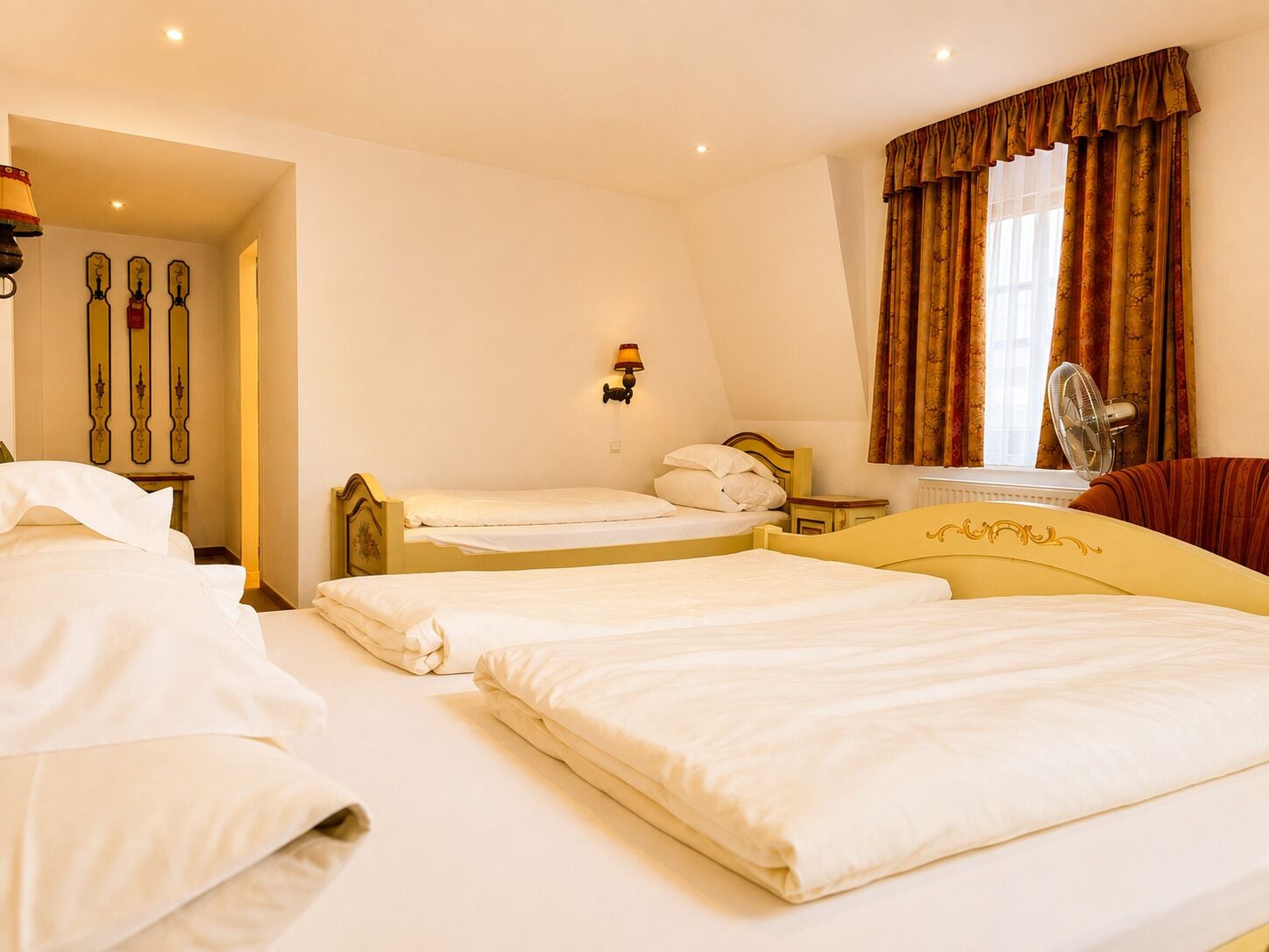 A hotel room with two beds, a sitting area with a sofa, a window with curtains, and a wall-mounted fan.