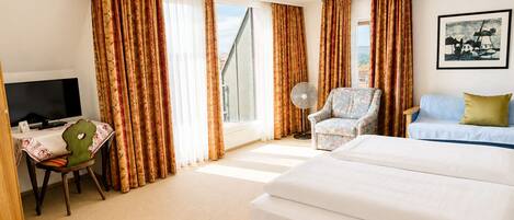 Habitación de hotel con cama, escritorio con televisor, una silla, un sofá y ventana con cortinas.