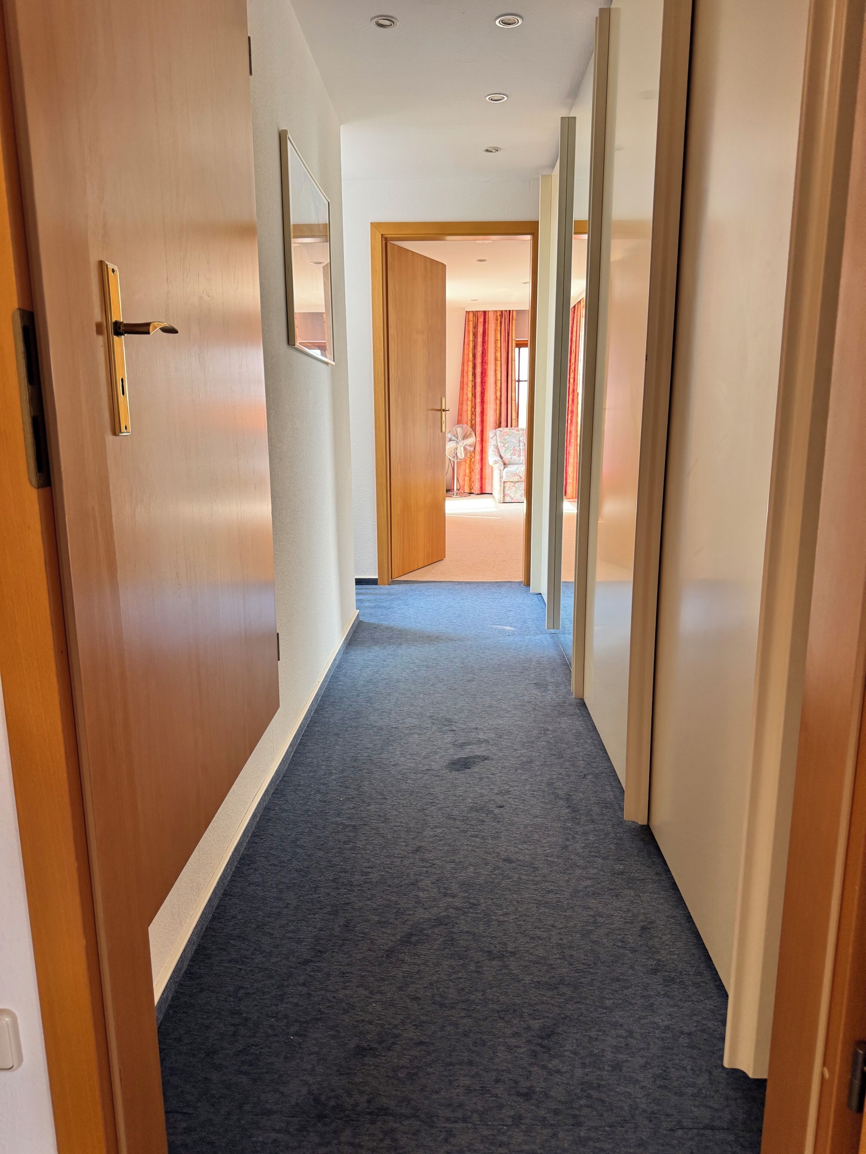 Een gang met tapijttegels, houten deuren en zicht op een kamer met een stoel en gordijnen.