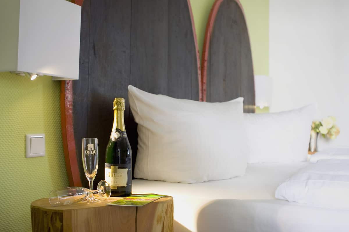 Une chambre d’hôtel avec un lit, une table de chevet en bois sur laquelle se trouvent une bouteille de champagne et un verre, et un mur au décor texturé.