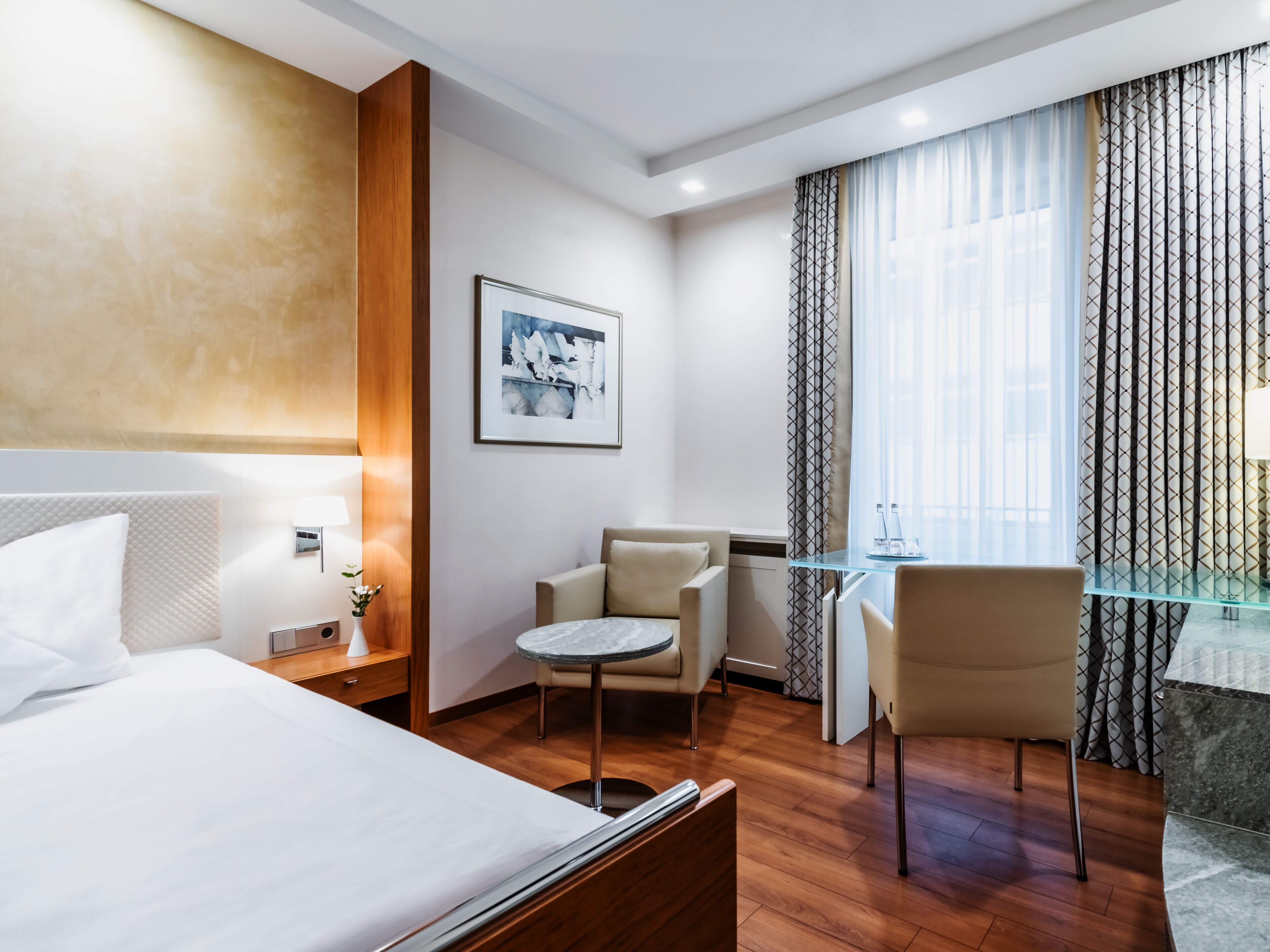 Una habitación de hotel con una cama, un escritorio, dos sillas, una mesita y un cuadro en la pared.