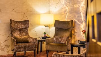 A corner room with a marble wall, two patterned armchairs, a small table with a lamp, and a carpeted floor.