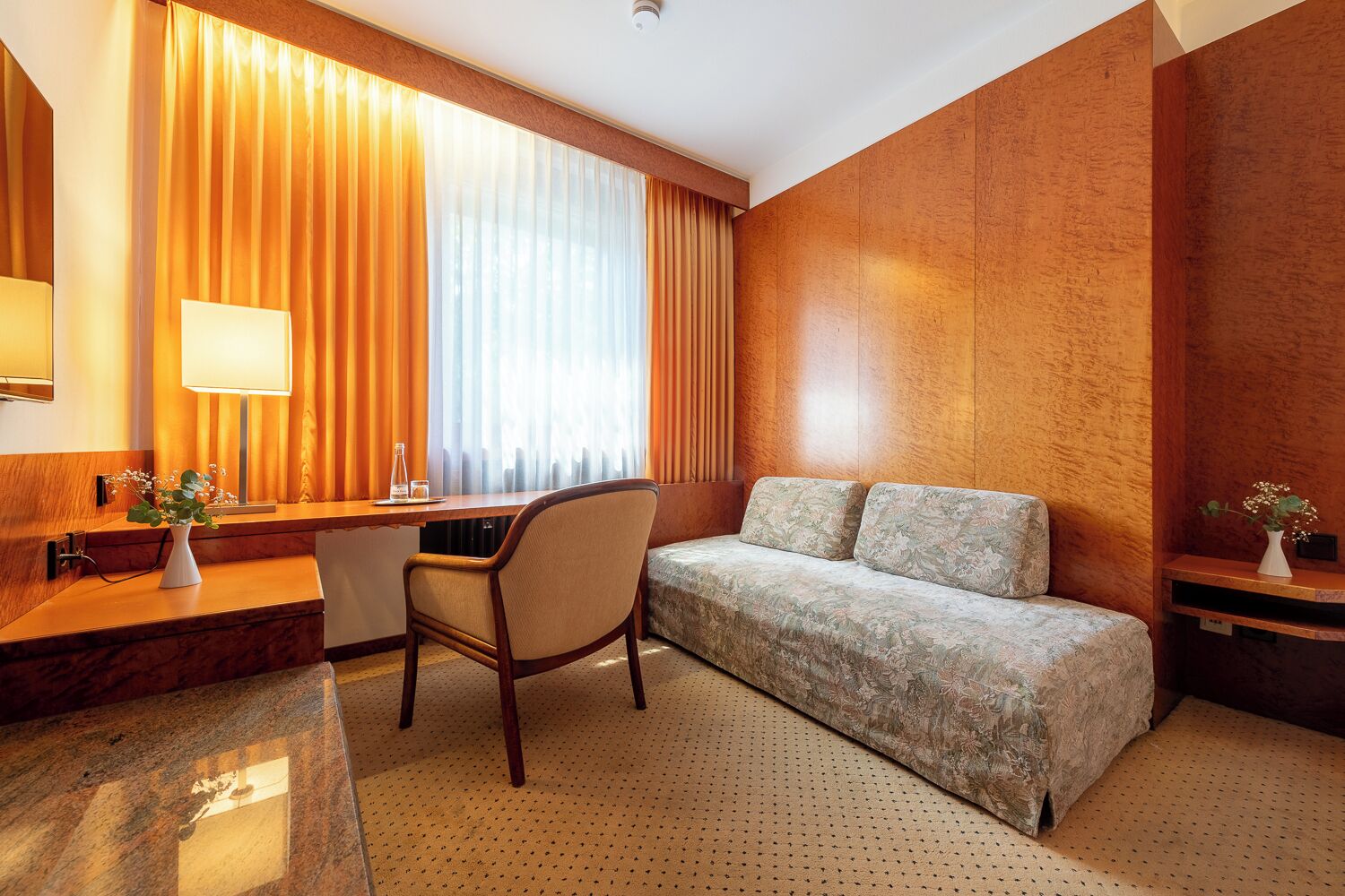 Habitación de hotel con pared de acento de madera, un escritorio con silla, sofá y un ventanal con cortinas.