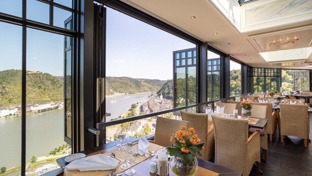 Un restaurant offrant une vue sur une rivière et des collines, doté de grandes fenêtres et de meubles en osier.