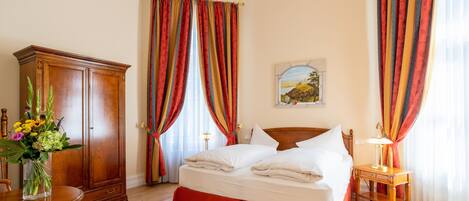 A hotel room with a wooden bed, a wooden wardrobe, a dining table with chairs, and a vase of flowers.