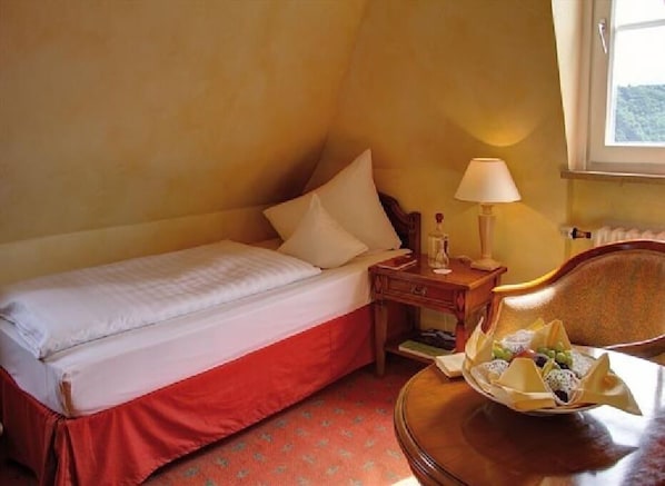 A hotel room with a single bed, a bedside table with a lamp, a chair, and a fruit basket. - Hotel Schloss Rheinfels (Sankt Goar)