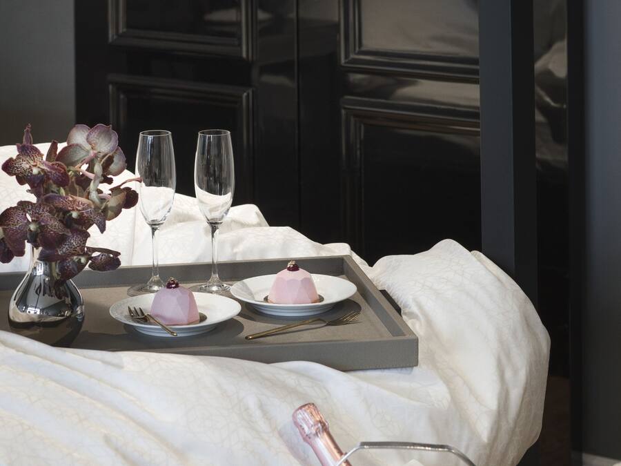 Una cama bien hecha con una bandeja de comida, un jarrón de flores y un cubo para champán.