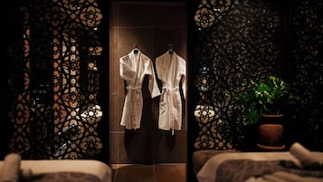 Habitación de hotel con dos camas, una planta en maceta y una puerta con dos batas blancas colgadas en ganchos.