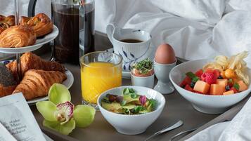 A breakfast tray with croissants, a coffee pot, a glass of orange juice, a bowl of fruit salad, and a cup of coffee.