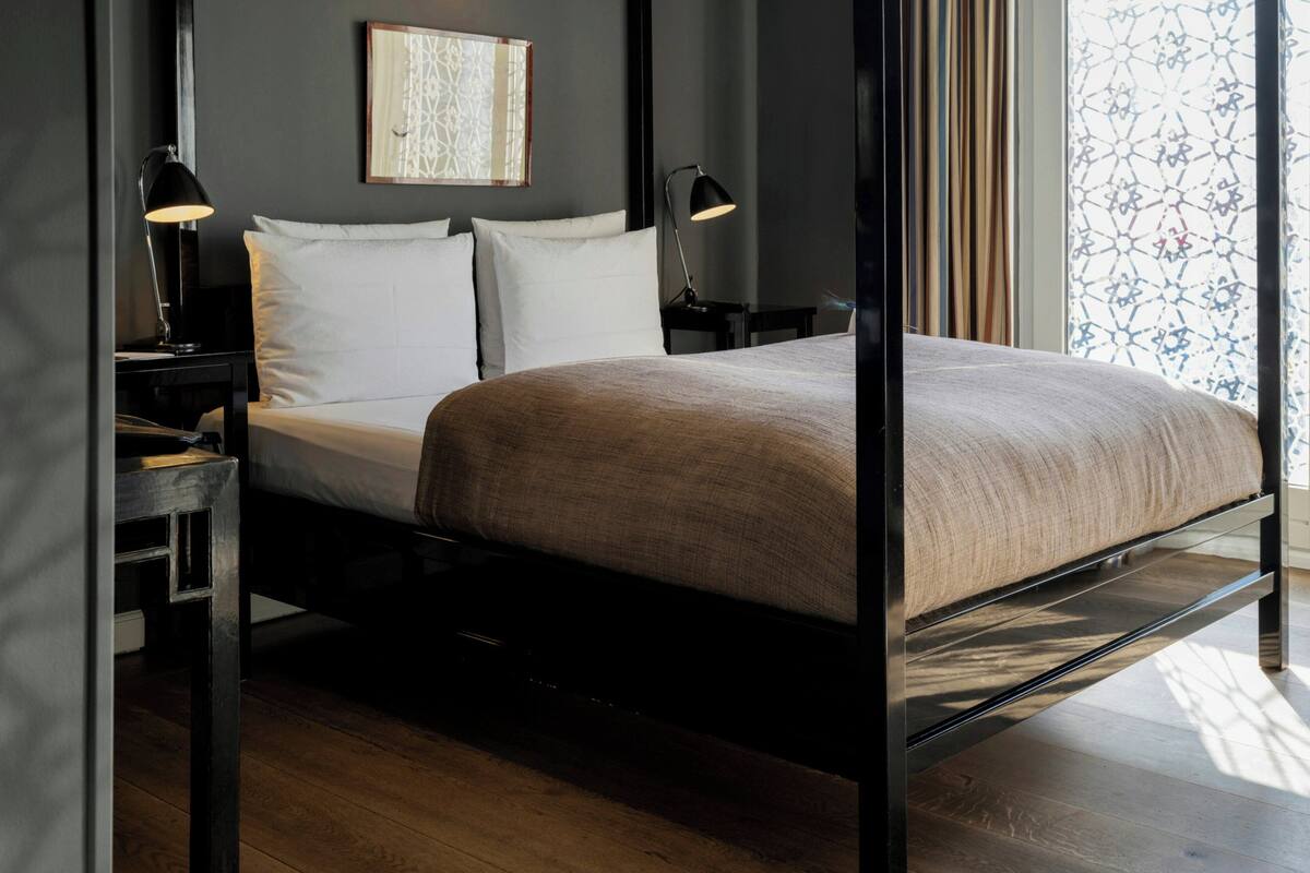 A modern bed with a metal frame, two bedside tables with lamps, a wall art piece, and a window with patterned curtains.
