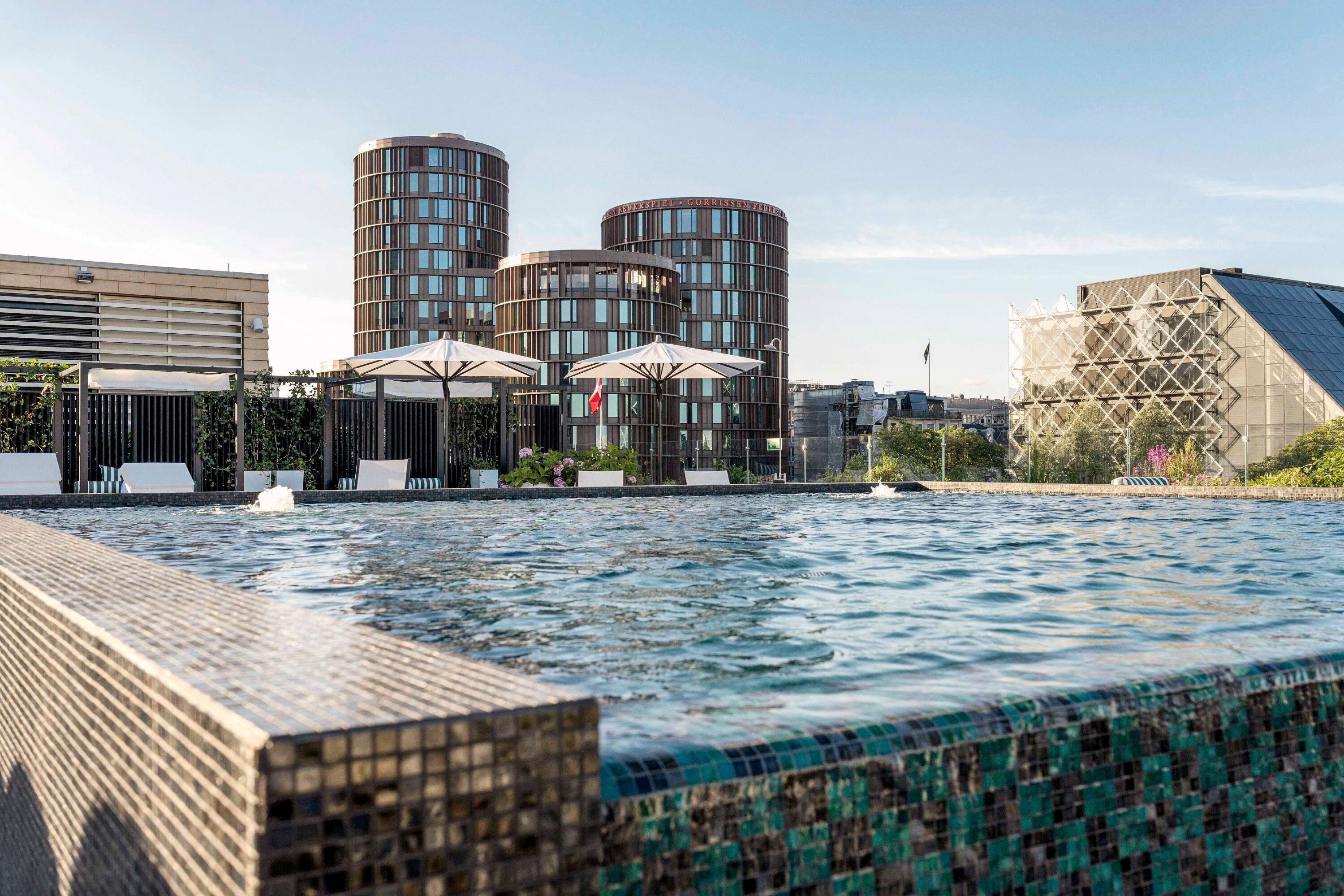 Una piscina con borde de azulejos, rodeada de edificios modernos y zonas de estar al aire libre.