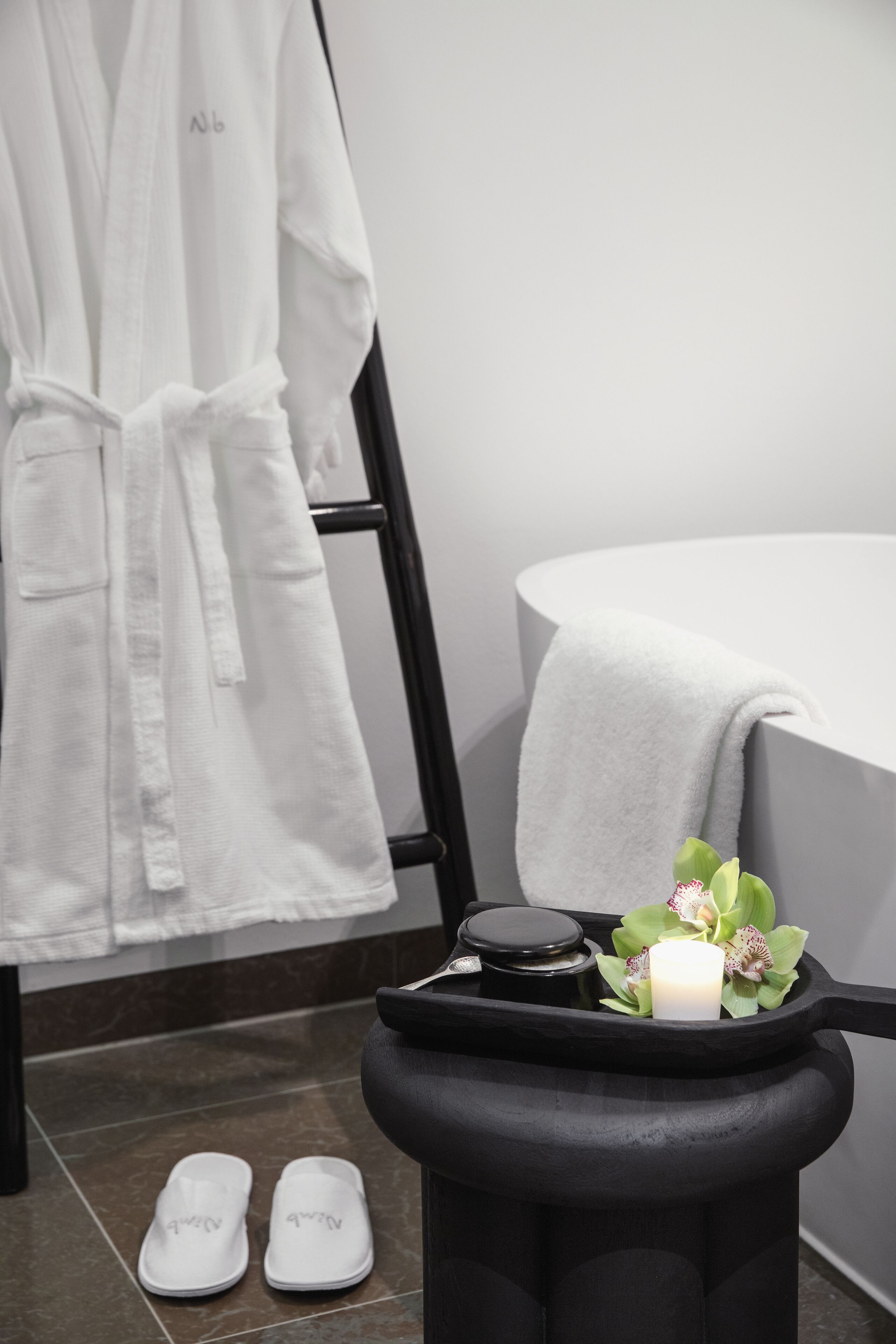 Un baño con una bañera, un perchero con una bata blanca, un taburete negro con una vela y flores, y un par de pantuflas en el suelo.