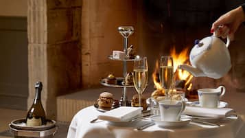 A table set for tea service with a teapot pouring, surrounded by a variety of pastries and a bottle of wine, with a fireplace in the background.