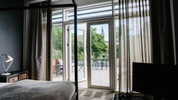 A hotel room with a bed, a desk with a lamp, a TV, and a sliding glass door leading to an outdoor patio with a table and chairs.