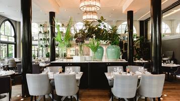 Ein Restaurant mit einer zentralen Insellounge, schwarzen Säulen und einer Kristallleuchter. Die Tische sind mit weißen Tischdecken und Stühlen gedeckt.