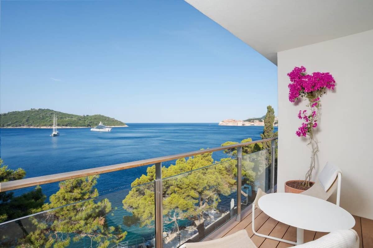 A balcony with a clear view of the sea, a white table, and a potted plant with pink flowers.