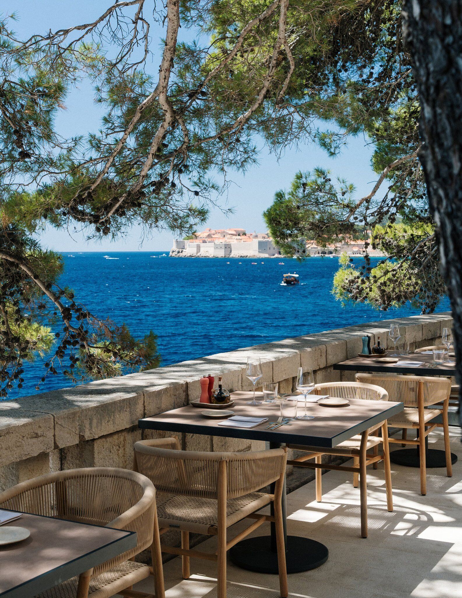 Un espace de restauration en plein air avec des tables en pierre et des chaises en osier, offrant une vue sur une mer d’un bleu profond et sur un bâtiment historique en arrière-plan.