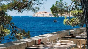 Un espace de restauration en plein air avec des tables en pierre et des chaises en osier, offrant une vue sur une mer d’un bleu profond et sur un bâtiment historique en arrière-plan.