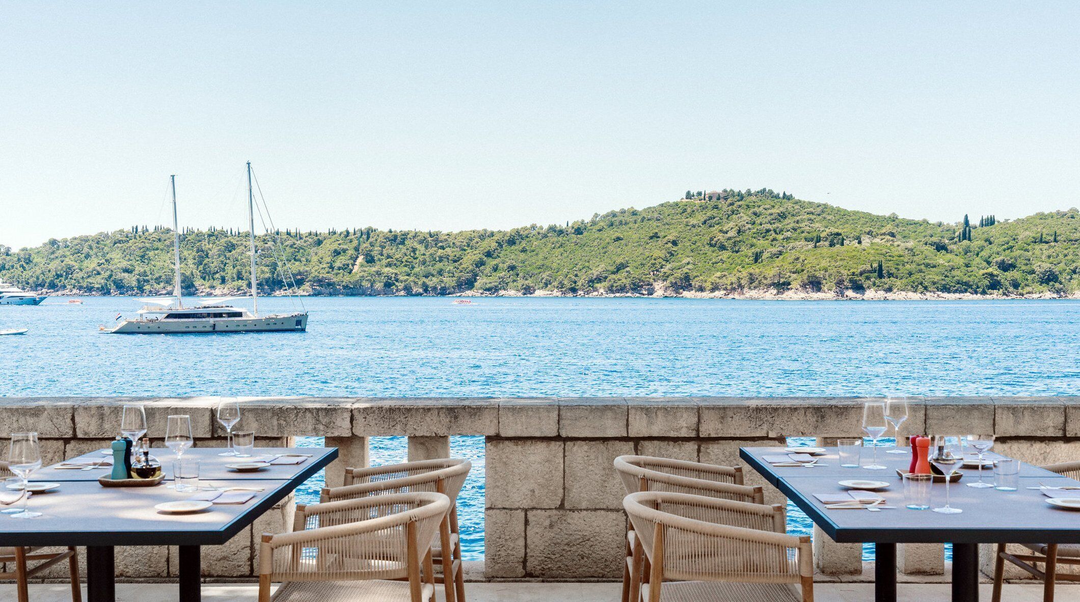 A seaside restaurant with tables and chairs facing a body of water, a yacht in the distance, and a hill with trees in the background.