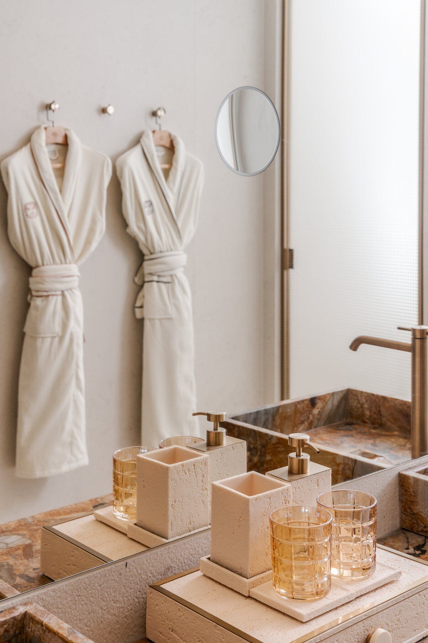 A bathroom with two white robes hanging on hooks, a mirror, and a marble sink with two glasses and a soap dispenser.