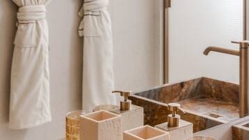 A bathroom with two white robes hanging on hooks, a mirror, and a marble sink with two glasses and a soap dispenser.