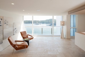 A modern hotel room with large windows, a view of the water, and two leather chairs.