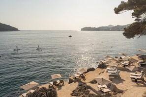A beach with sun loungers and umbrellas, a rocky shoreline, and boats on the water.
