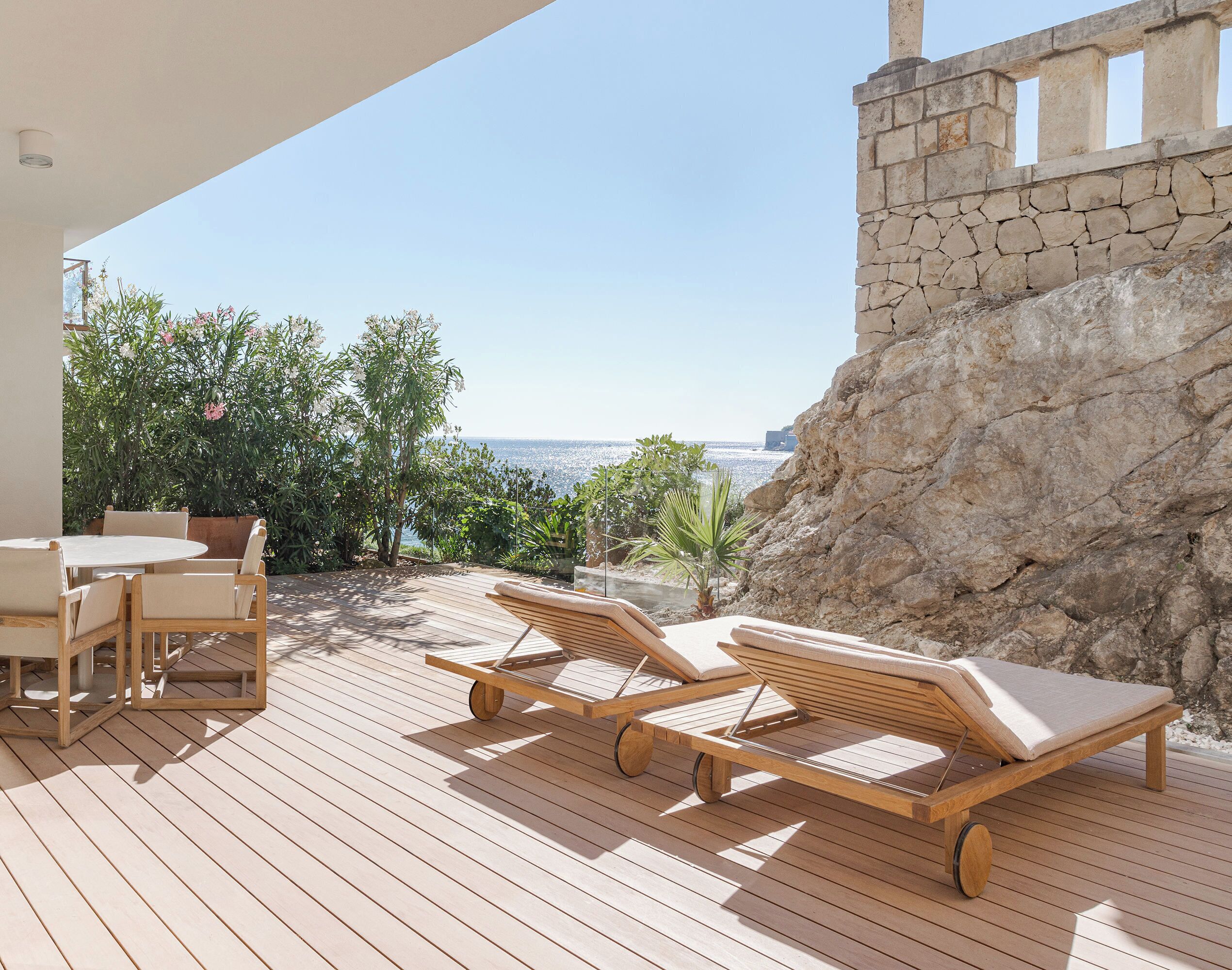 Um deck de madeira com cadeiras de descanso e uma mesa, com vista para um penhasco rochoso e o mar.