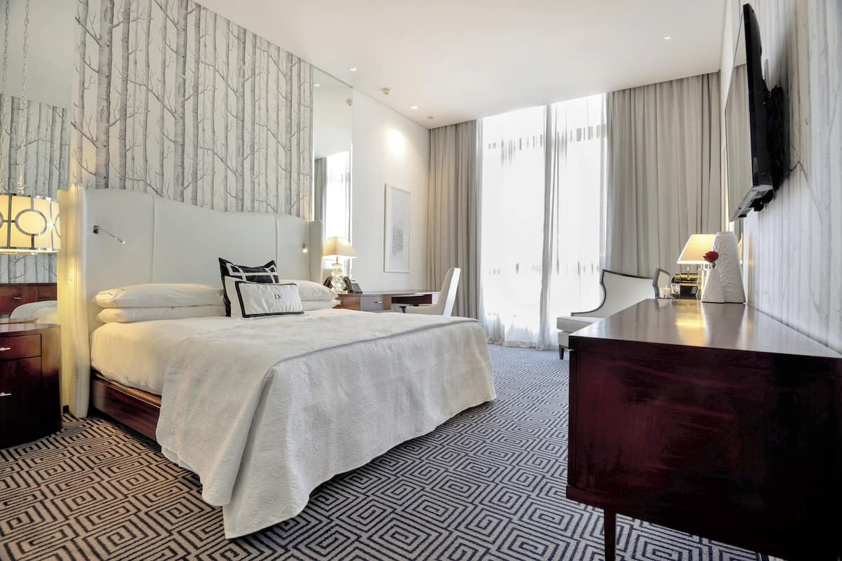A modern hotel room with a large bed, a desk, a chair, a TV, and a patterned carpet.