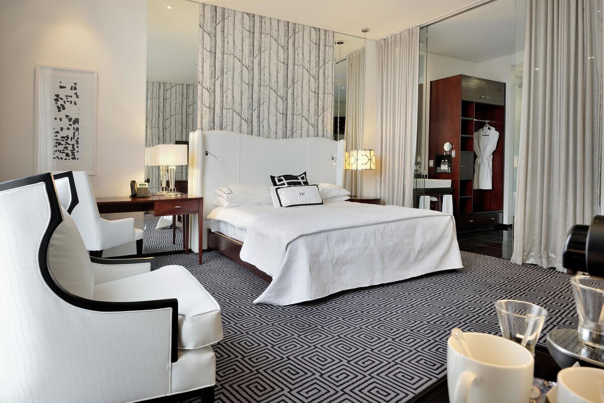 A hotel room with a large bed, a white armchair, a desk with a lamp, and a wardrobe.
